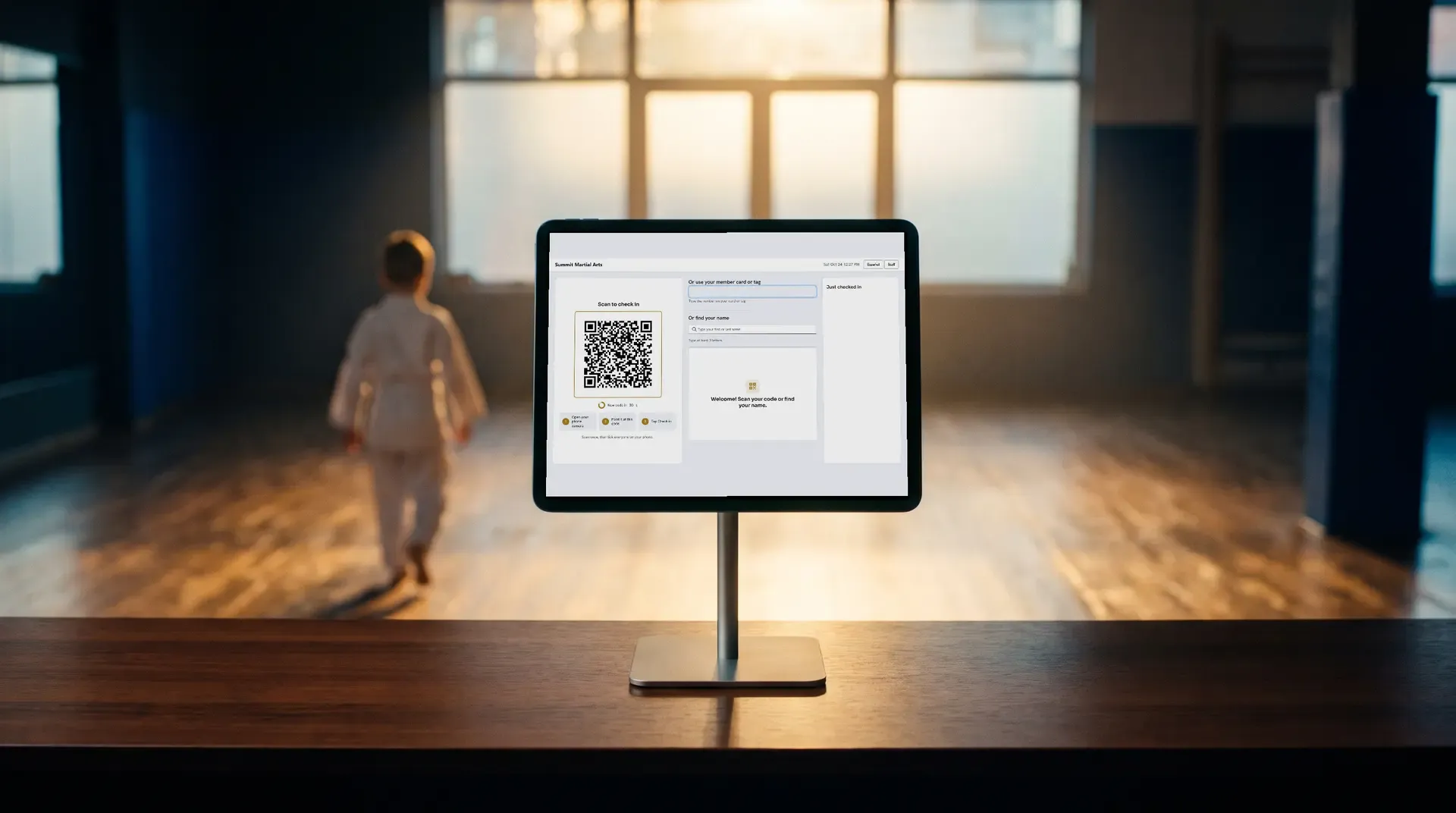 A tablet on a front desk showing the check-in screen with a QR code, a card number box and a name search, a student walking into the training hall behind it.